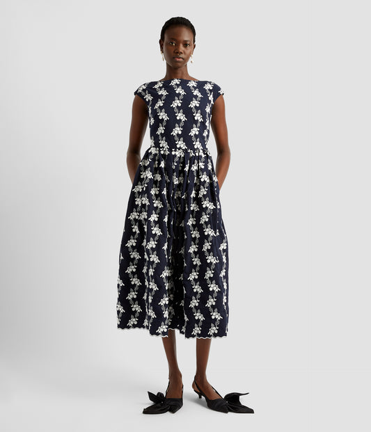 A classic 1950s fit and flare dress. The hem midi dress features a boatneck, cap sleeves and large flared skirt. The scalloped hem midi dress is made from embroidered navy fabric with ditsy white floral embroidery.  