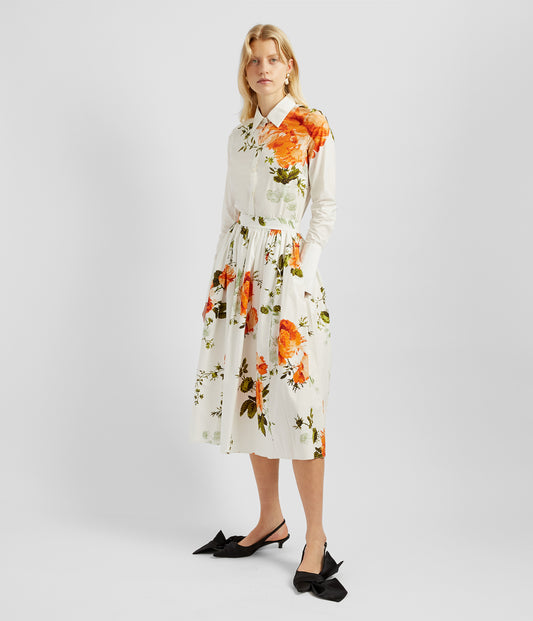 A cotton poplin white midi skirt with a fitted waist and voluminous silhouette. The skirt has a rose print in orange and green and features relaxed hidden pockets.