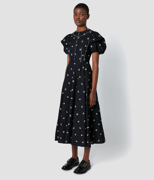 Image of a model wearing a jacquard midi dress in a classic navy with a delicate white floral print. The dress features a defined waist, full skirt and puff sleeves. 