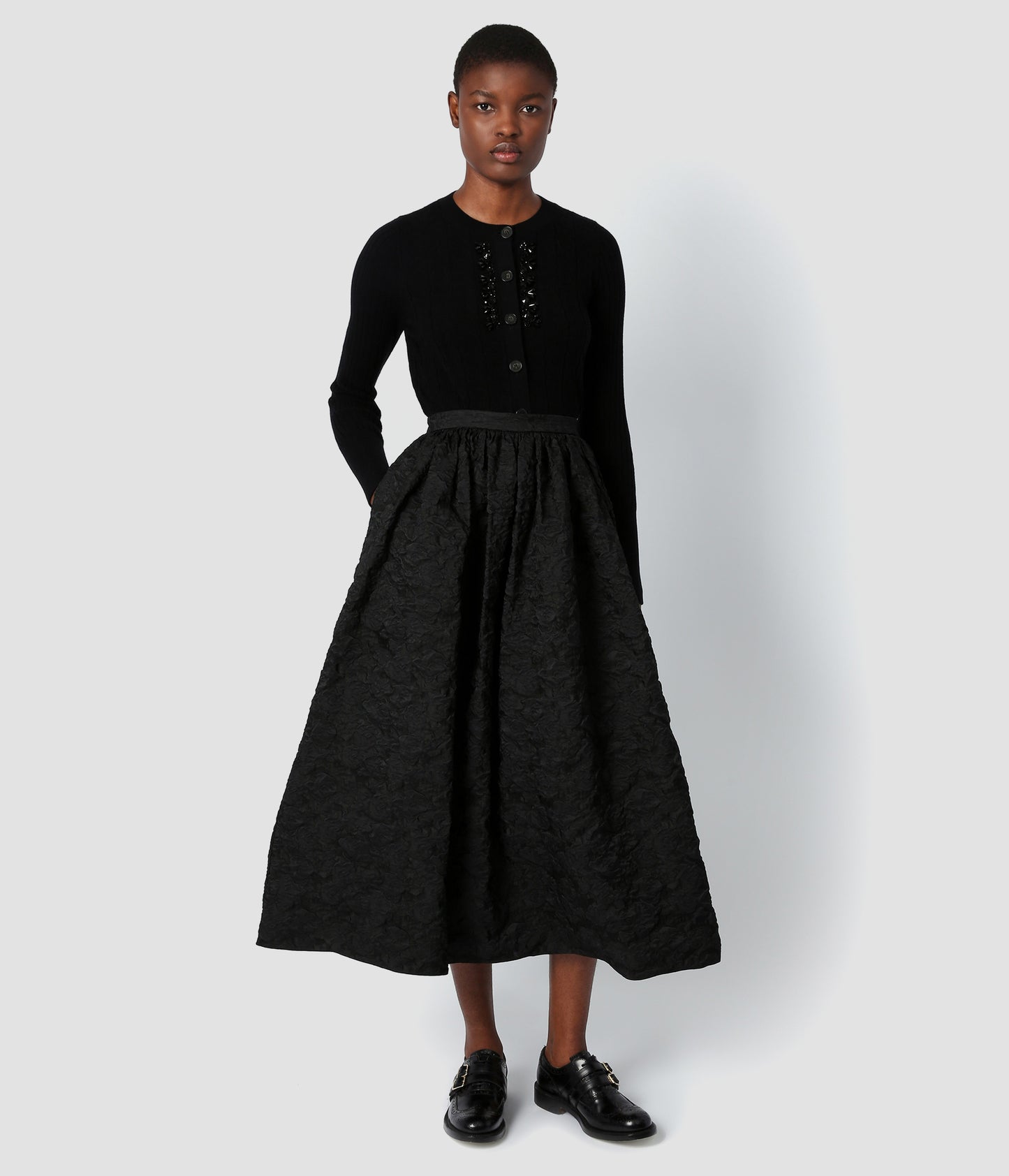 Black voluminous skirt made from structured cloque that is beautifully textured. The skirt is worn by a model who is using the pockets. The voluminous skirt is mid length, falling above the ankle.