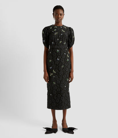 A black crew neck midi dress in floral matelassé with crystal and beeded insect details. The midi dress features a crew neck bodice, pencil skirt and tulip sleeves.