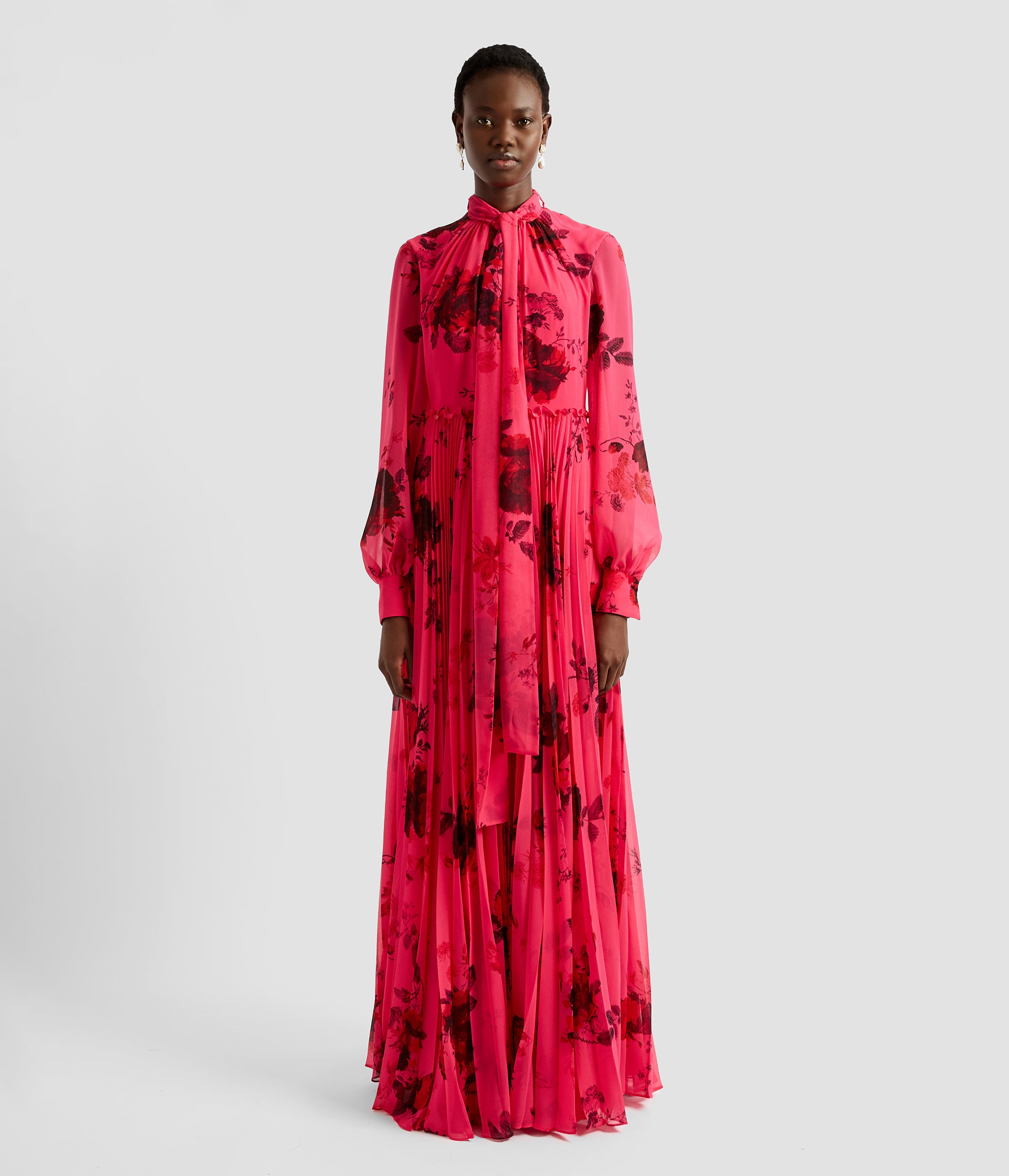 This vibrant blouson gown with sleeves is made from pleated voile which creates a stunning sense of volume. The fabric is bright cerise and features a dark floral. The sleeves are full length and the neck features a tie closure. 