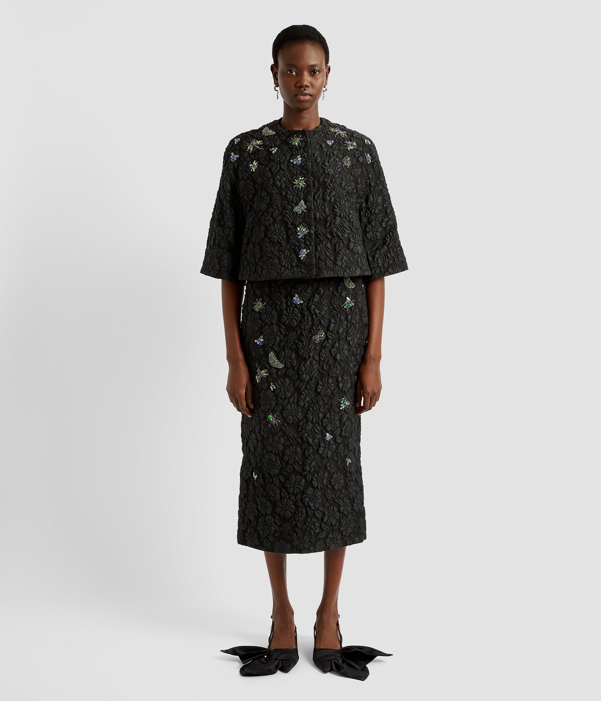 A black cropped cape by desinger Erdem in a textured floral fabric. The cropped cape has elbow length sleeves and crystall embellished insect embroidery and buttons. 