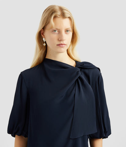 A silk draped blouse in navy by designer Erdem. The draped blouse has elbow length with gathered sleeves and a tie neck line that drapes down the front. 